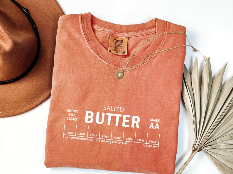 May include: A coral-coloured t-shirt with the text "SALTED BUTTER AA" printed in white. The shirt also includes a measurement guide for tablespoons and cups of butter.