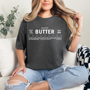 May include: A woman wearing a dark grey t-shirt with the text "SALTED BUTTER GRADE AA" and a measurement chart. The shirt is a relaxed fit and has a distressed look.