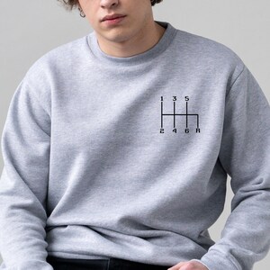 May include: A grey sweatshirt featuring a black graphic of a 6-speed manual transmission gear shift pattern. The numbers 1, 2, 3, 4, 5, and 6 are printed on the gear shift pattern, along with the letter "R" for reverse.