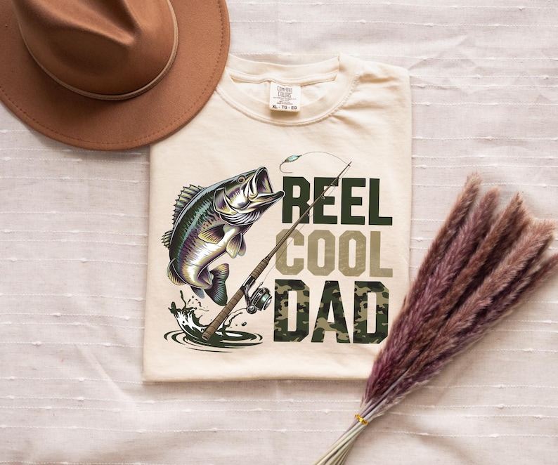 May include: Cream-colored t-shirt featuring a leaping fish graphic with a fishing rod. The text reads "REEL COOL DAD" in green and camouflage lettering. A brown hat is visible in the upper left corner.