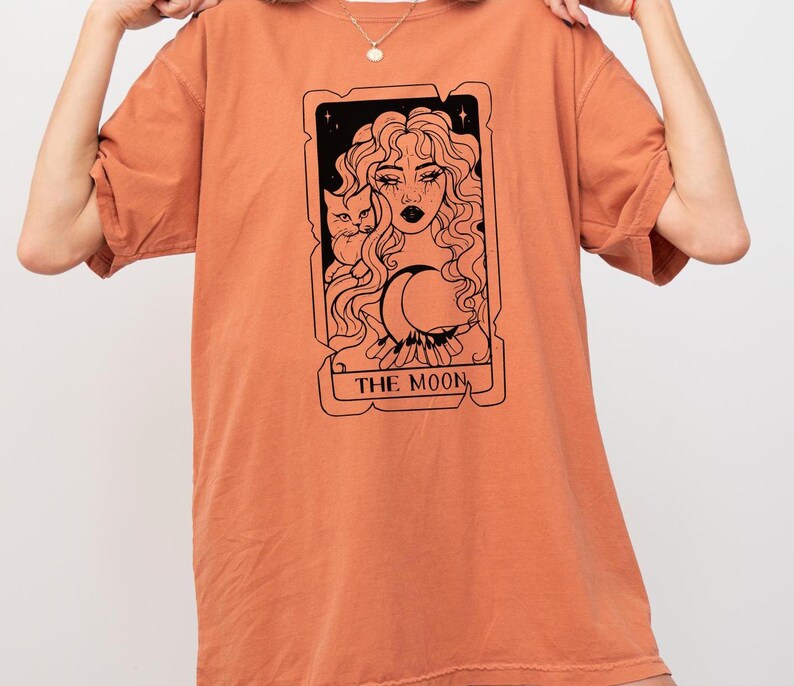 May include: A burnt orange t-shirt with a black and white illustration of a tarot card depicting "The Moon". The card features a woman with long hair holding a crescent moon and a cat.
