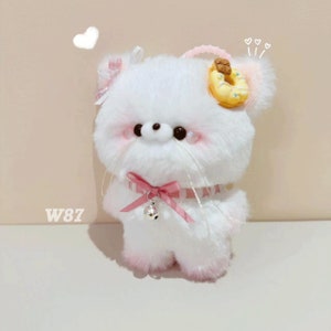May include: White plush toy with pink accents, a pink bow, and a donut on its head. The toy has a bell on its neck and is standing on a white surface. W87