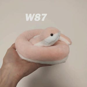May include: A plush toy shaped like a coiled snake with soft light pink fur on the top and white fur on the underside. The snake has a small black eye and a white snout. The text W87 is visible in white above the plush snake toy.