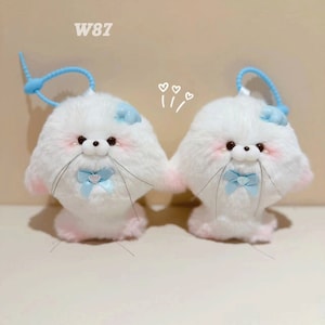 Può includere: Due peluche di foca bianche con fiocchi blu e dettagli di fiori blu. I peluche hanno guance rosa e occhi neri. Sono attaccati a un elastico blu.