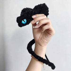 Puede incluir: Un accesorio de serpiente de felpa negra con un ojo azul brillante y una pupila negra vertical. El material texturizado y difuso cubre toda la longitud de la serpiente, que incluye una sección de cola en espiral. El texto W87 es visible en el cuerpo.