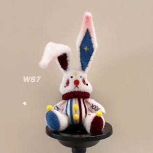 Puede incluir: Un conejo de peluche blanco con orejas largas, adornado con detalles azules, rojos y amarillos. El conejo tiene un diseño caprichoso, una nariz roja y una bufanda a rayas. El texto "W87" es visible en el lado izquierdo de la imagen.