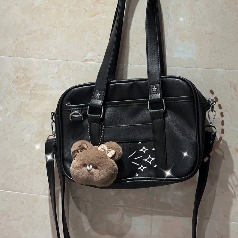 May include: Black faux leather satchel bag with a brown plush bear charm and white star accents. The bag has a top handle and a long adjustable shoulder strap.