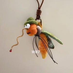May include: A plush insect ornament featuring an orange head with white googly eyes, a green crocheted hat, and a long, curly tan proboscis ending in a red bead. The body is orange with yellow stitching, thin black wire legs, and green wings.
