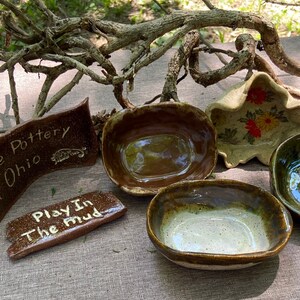May include: Handmade pottery bowls in various colors and designs, including a brown bowl with a speckled glaze, a green bowl with a smooth glaze, and a white bowl with a floral design. The bowls are arranged on a brown surface with a wooden branch in the background. The text "Handmade Clayton Ohio Pottery" and "Play In The Mud" are visible on two of the bowls.