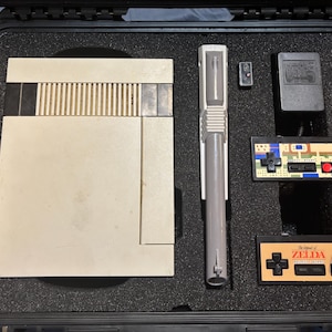Op de afbeelding: Een vintage Nintendo Entertainment System (NES) console, lichtgrijs, met een bijpassende lichtgrijze controller en twee gamecontrollers, allemaal in een zwarte koffer. De controllers zijn voorzien van het label "The Legend of Zelda".
