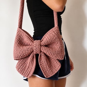 Può includere: Una borsa a tracolla all'uncinetto color rosa antico a forma di grande fiocco. La borsa presenta una struttura in filato testurizzato cucito a mano con un'ampia tracolla e un dettaglio a nodo centrale, creando una sagoma a fiocco.