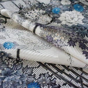May include: Close-up of a folded fabric with a floral pattern. The design features large white and gray flowers with blue accents, set against a cream background. The fabric has a textured appearance.