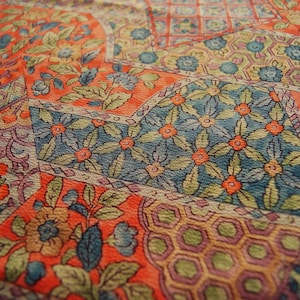 Può includere: Primo piano di un tessuto con un design patchwork. Il tessuto presenta un vivace mix di colori, tra cui arancione, blu, verde e viola. Sono visibili motivi floreali e geometrici, che creano una trama visiva ricca e dettagliata.