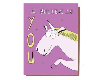 May include: A greeting card with a purple background and a cartoon unicorn illustration. The text "I BELIEVE IN YOU" is written in white and yellow. The unicorn is pink with a white horn and yellow mane. The card is on a brown background.