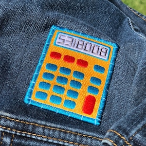 May include: A denim jacket with a colorful embroidered patch of a calculator. The calculator patch is yellow with red, blue, and white details, and the display reads "5318008". The jacket is a medium wash denim.