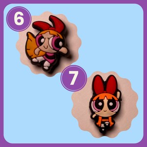 May include: Two cartoon character charms, one with orange hair and a red bow, the other with blonde hair and a yellow bow. Both characters have large eyes and are wearing pink dresses. The charms are numbered 6 and 7.