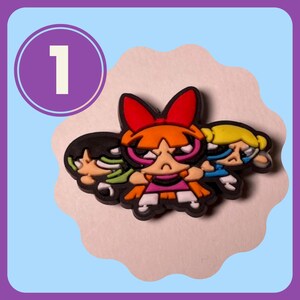 May include: A set of three cartoon characters from the Powerpuff Girls show. The characters are Blossom, Bubbles, and Buttercup. Blossom is in the center and has red hair, a red bow, and a pink dress. Bubbles is on the right and has blonde hair and a blue dress. Buttercup is on the left and has black hair and a green dress.