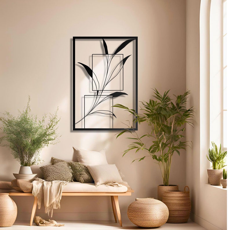 Op de afbeelding: Zwarte metalen wandkunst met een gestileerde tak met bladeren in een vierkant frame. Het frame is geplaatst tegen een lichtbeige muur.