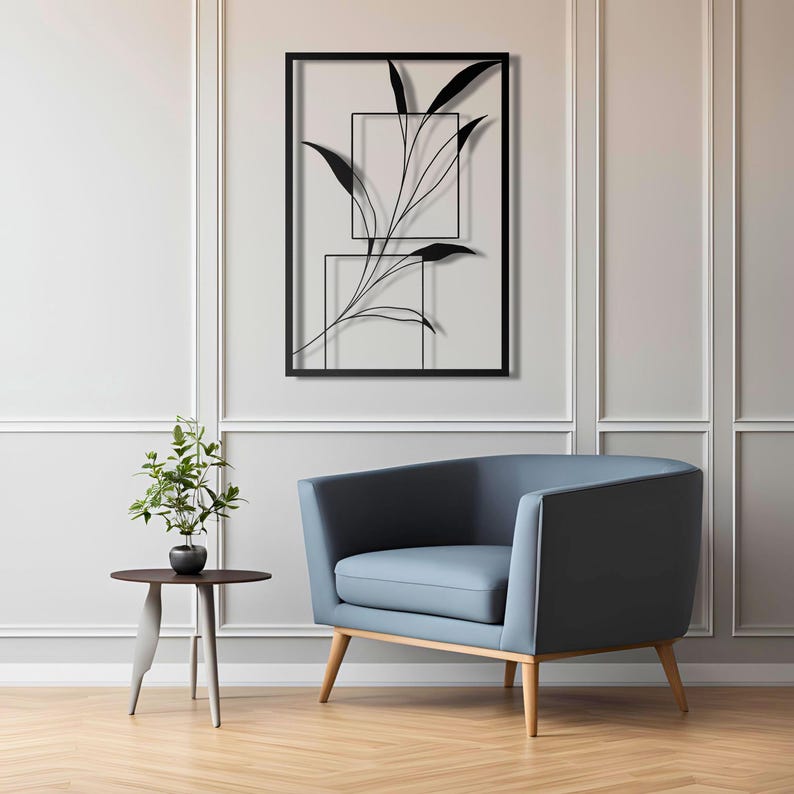 Op de afbeelding: Zwarte metalen wandkunst met een gestileerde tak met bladeren in twee rechthoekige frames. De kunst hangt aan een witte muur boven een blauwe fauteuil.