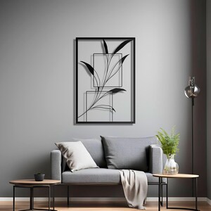 Op de afbeelding: Zwarte metalen wandkunst met een gestileerde tak met bladeren in een rechthoekig frame. Het kunstwerk wordt getoond op een grijze muur boven een grijze bank met kussens en een deken.