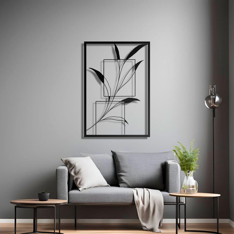 Op de afbeelding: Zwarte metalen wandkunst met een gestileerde tak met bladeren in een rechthoekig frame. Het kunstwerk wordt getoond op een grijze muur boven een grijze bank met kussens en een deken.
