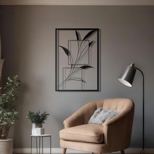 Op de afbeelding: Zwarte metalen wandkunst met een gestileerde tak met bladeren in een vierkant frame. De kunst hangt aan een grijze muur boven een bruine fauteuil met een grijs kussen.