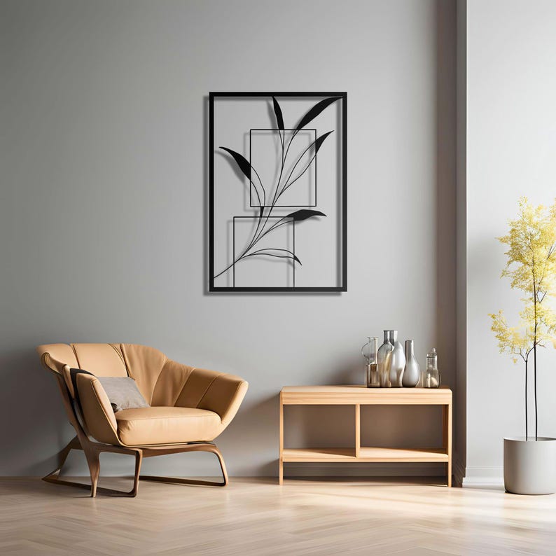 Op de afbeelding: Zwarte metalen wandkunst met een gestileerde tak met bladeren in twee vierkante frames. De kunst hangt aan een grijze muur boven een lichtbruin houten plank met twee planken.