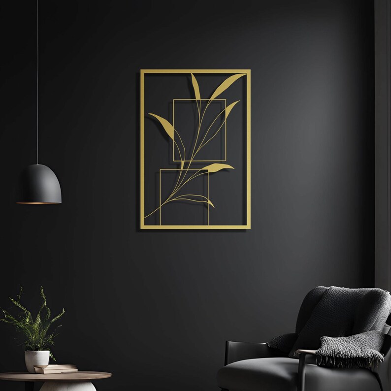 Op de afbeelding: Gouden metalen wandkunst met een gestileerde tak met bladeren. De kunst is ingelijst in een vierkante vorm en heeft een minimalistisch ontwerp.