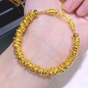 Pu&ograve; includere: Un braccialetto color oro con un design strutturato e perlato. Il braccialetto &egrave; composto da piccole perle irregolari infilate insieme, creando un accessorio unico. La chiusura &egrave; visibile e il braccialetto &egrave; mostrato sul polso di una persona.