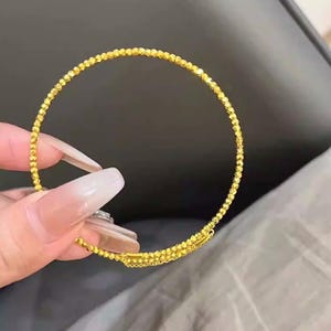 May include: A gold bracelet with a beaded design. The bracelet is made of small, faceted beads that catch the light, creating a sparkling effect. The clasp is a simple bar and loop closure.