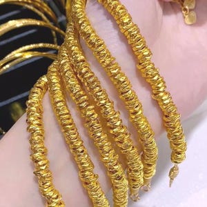Pu&ograve; includere: Una collezione di braccialetti color oro con un design strutturato e scanalato. Diversi braccialetti sono impilati, mostrando i dettagli intricati e la lucentezza dei gioielli. I braccialetti sono esposti su uno sfondo chiaro.