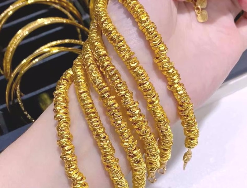 Pu&ograve; includere: Una collezione di braccialetti color oro con un design strutturato e scanalato. Diversi braccialetti sono impilati, mostrando i dettagli intricati e la lucentezza dei gioielli. I braccialetti sono esposti su uno sfondo chiaro.