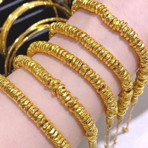 Pu&ograve; includere: Primo piano di diversi braccialetti color oro con un design strutturato e scanalato. I braccialetti sono indossati al polso di una persona, evidenziando il loro aspetto luminoso e lucente. Altri braccialetti sono visibili sullo sfondo, suggerendo una collezione di gioielli.