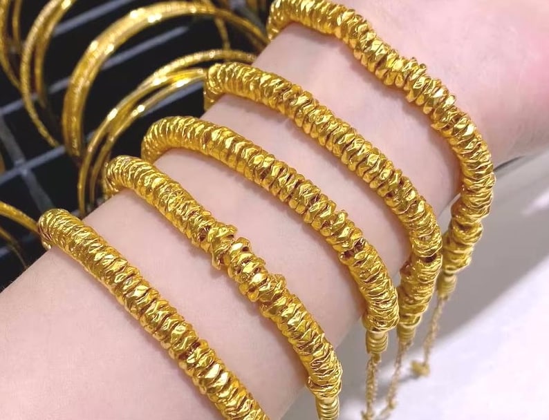 Pu&ograve; includere: Primo piano di diversi braccialetti color oro con un design strutturato e scanalato. I braccialetti sono indossati al polso di una persona, evidenziando il loro aspetto luminoso e lucente. Altri braccialetti sono visibili sullo sfondo, suggerendo una collezione di gioielli.