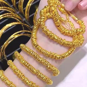 Pu&ograve; includere: Primo piano di gioielli in oro, tra cui braccialetti strutturati e una collana a corda intrecciata. Diversi braccialetti d'oro sono visibili anche sullo sfondo.