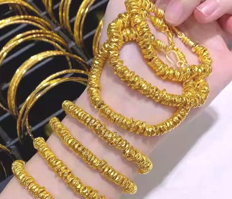 Pu&ograve; includere: Primo piano di gioielli in oro, tra cui braccialetti strutturati e una collana a corda intrecciata. Diversi braccialetti d'oro sono visibili anche sullo sfondo.