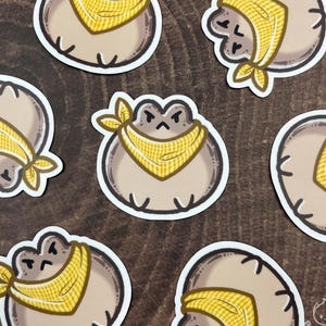 Puede incluir: Una colección de pegatinas con una rana de dibujos animados con un pañuelo amarillo. La rana es marrón con un contorno blanco. El pañuelo tiene un estampado de cuadros amarillos y blancos. Las pegatinas están dispuestas sobre una superficie de madera.