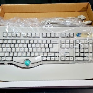 May include: A light gray computer keyboard with a built-in trackball and wrist rest, packaged in a cardboard box. The keyboard has white keys and a teal trackball. The brand name "ECO" is visible on the top right corner.