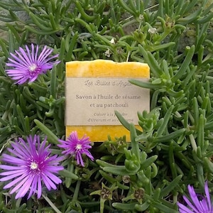 Op de afbeelding: Een gele zeep met een bruin label waarop staat "Les Bulles d'Ange", "Savon à l'huile de sésame et au patchouli", "Coloré à la poudre d'Urucum et au curcuma". De zeep ligt op een bed van groene planten met paarse bloemen.
