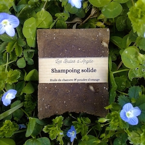 Può includere: Una barra di shampoo solido marrone con un'etichetta che recita "Les Bulles d'Angèle Shampooing solide Huile de chanvre & poudre d'orange" circondata da foglie verdi e fiori blu.