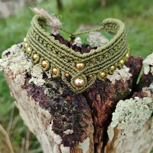 Peut inclure: Collier ras du cou macramé vert olive avec des perles dorées. Le collier fait main est présenté sur une surface en bois vieilli, mettant en valeur les nœuds et les perles. Un accessoire de style bohème.
