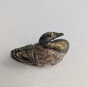 Op de afbeelding: Een kleine, decoratieve eendenfiguur. De eend heeft een gedetailleerd, getextureerd lichaam met een gespikkeld patroon in bruin- en beigetinten. De kop en snavel zijn donkergrijs met gouden accenten, de vleugels groen en goud.