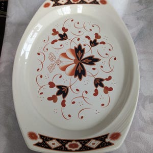May include: Oval serving platter with a cream-colored base. The platter features a floral design in shades of orange, black, and brown. The rim has a decorative border with geometric and floral elements. A vintage piece for serving food.