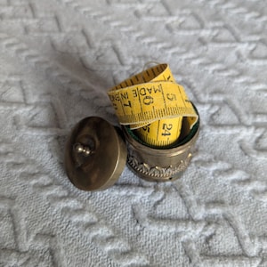 May include: A small, round, antique-style brass container with a removable lid, featuring a decorative embossed border. Inside the container sits a coiled yellow measuring tape with black markings, clearly displaying the text MADE IN ENGLAND.