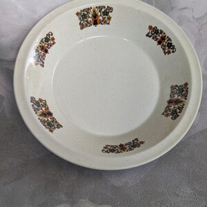May include: A round, off-white ceramic plate with a speckled surface. The plate features decorative floral patterns in shades of brown, orange, and green around the rim. The plate is designed for serving food.
