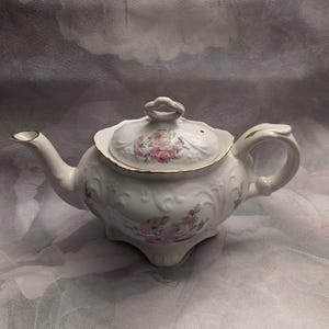 Pode incluir: Bule de porcelana branca com um design floral com rosas cor-de-rosa. A chaleira tem uma borda dourada e uma tampa com uma pega.