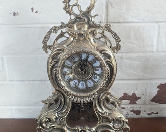 Op de afbeelding: Oude messing klok met een witte wijzerplaat en Romeinse cijfers. De klok heeft ingewikkeld krulwerk en een decoratieve bovenkant. Een vintage stuk, ideaal voor de inrichting van een huis. Meet ongeveer 20 cm hoog.