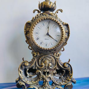 May include: An antique brass-colored mantel clock with a white clock face and Roman numerals. The clock has an ornate, decorative design with a detailed base and top finial. The clock's hands indicate the time.