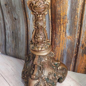 Può includere: Portacandele ornato in stile antico nei toni dell'oro e del bronzo. La base e lo stelo in metallo presentano intagli intricati e dettagliati. Il portacandele ha una finitura anticata, che gli conferisce un aspetto vintage. Altezza circa 30 cm.