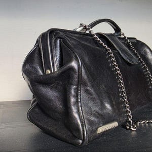 Könnte beinhalten: Eine schwarze Lederhandtasche im Doktortaschen-Stil mit strukturiertem Rahmenverschluss, einem Tragegriff und einem silberfarbenen Metallkettenschulterriemen. Die Tasche hat eine kleine rechteckige silberfarbene Metallplakette mit dem Namen VECCHI.